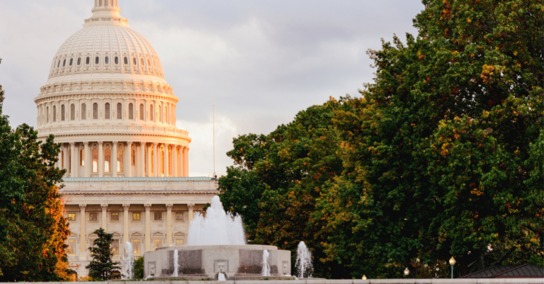 The U.S. Capitol building illuminated by warm light, partially framed by leafy green trees, with a fountain in the foreground under a cloudy sky.