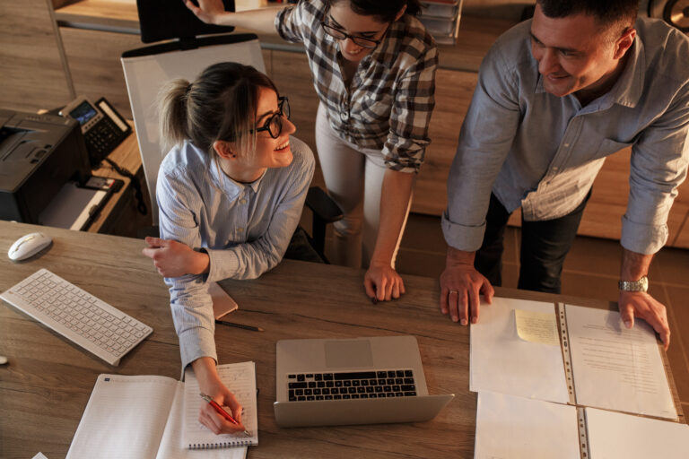 Three colleagues gathered around a large wooden desk in an office workspace. One person sits at the desk writing in a notebook beside an open laptop, while two others stand nearby leaning over documents spread across the table. A keyboard, computer mouse, printer, folders and notes are visible, creating a collaborative work environment with warm indoor lighting.