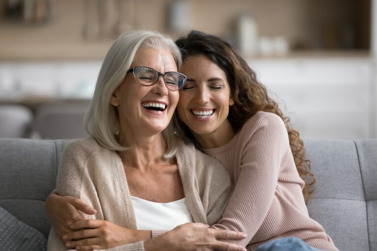 Close-up of a mother and daughter seated on a couch at home, leaning into each other with eyes closed and smiling, one wearing glasses with gray hair and the other with long curly hair and a light sweater, arms wrapped around each other in an embrace.