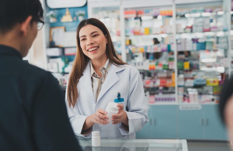 A pharmacist reviews a prescription medication with a customer at a community pharmacy counter. The image represents the exchange of prescription information and the communication between pharmacies, providers and health plans that supports safe, accurate medication management during the NCPDP F6 transition.