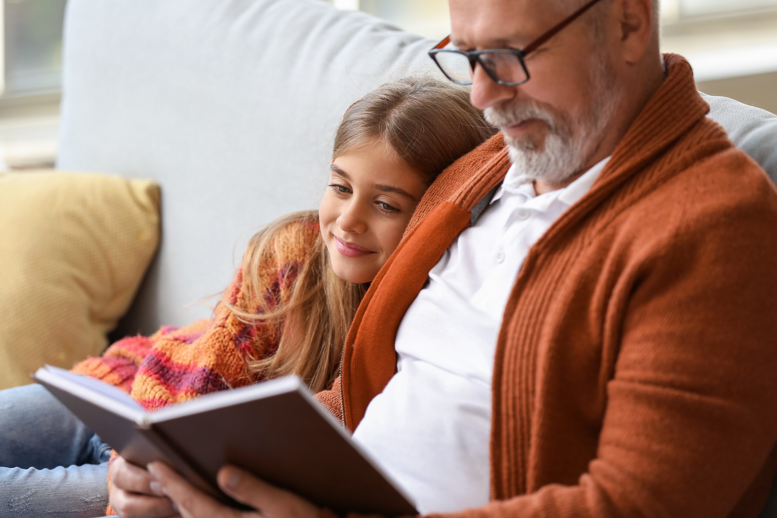Male wearing a white shirt and orange sweater sitting on a couch with a child leaning against them, holding a book. The image represents comfort and ease of mind.